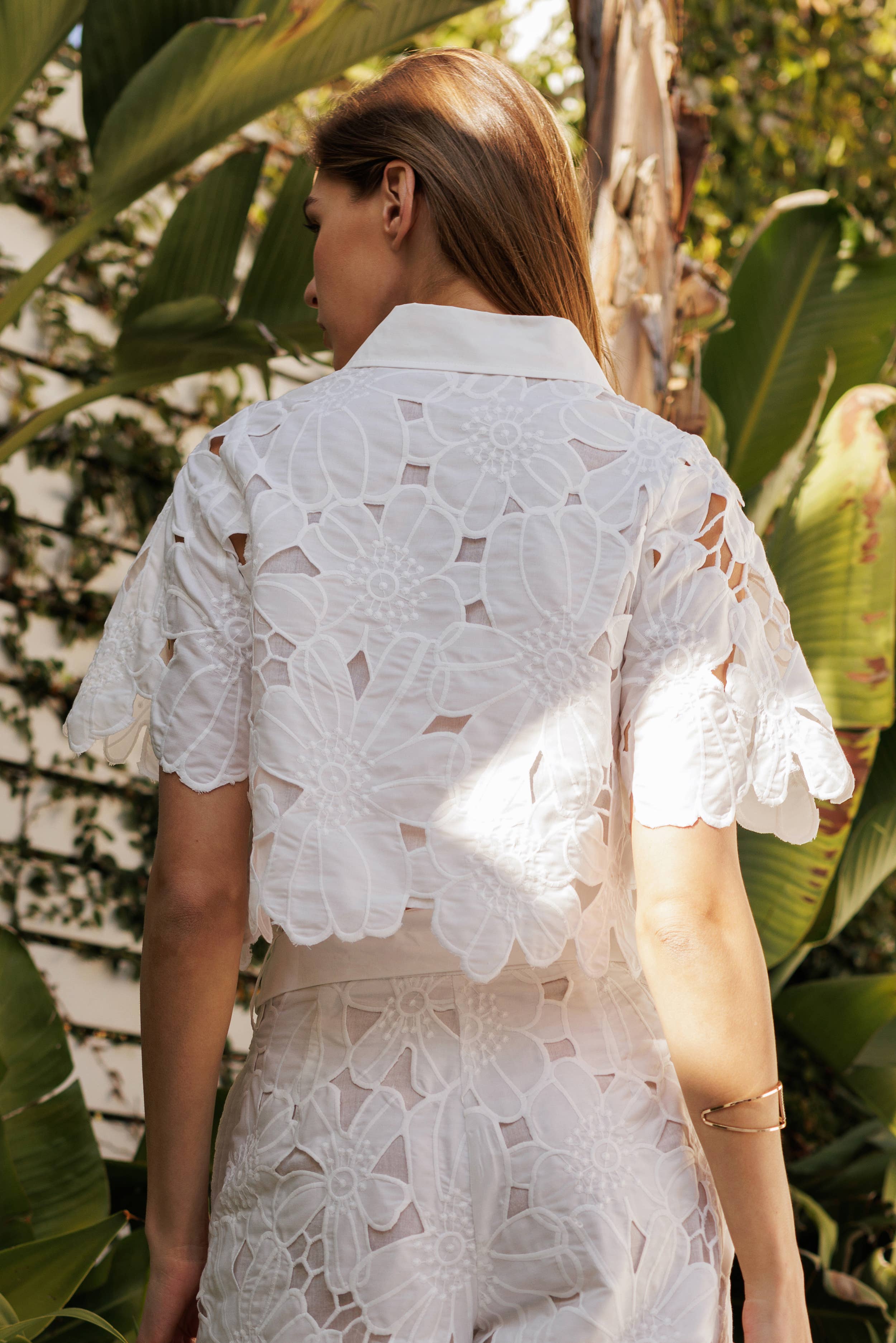 Back view of a woman wearing a white floral lace blouse among lush greenery, showcasing intricate lace details.