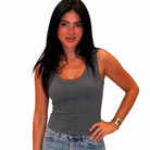 A woman with long black hair wearing a dark gray ribbed tank top and blue jeans, standing against a plain white background.