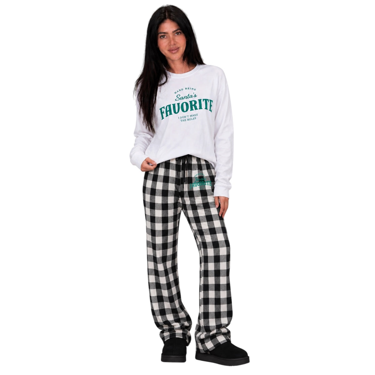 A woman modeling a white long-sleeve crewneck sweatshirt with green 'Santa’s Favorite' text on the chest, paired with black and white gingham plaid lounge pants and black slip-on shoes, against a plain white background.