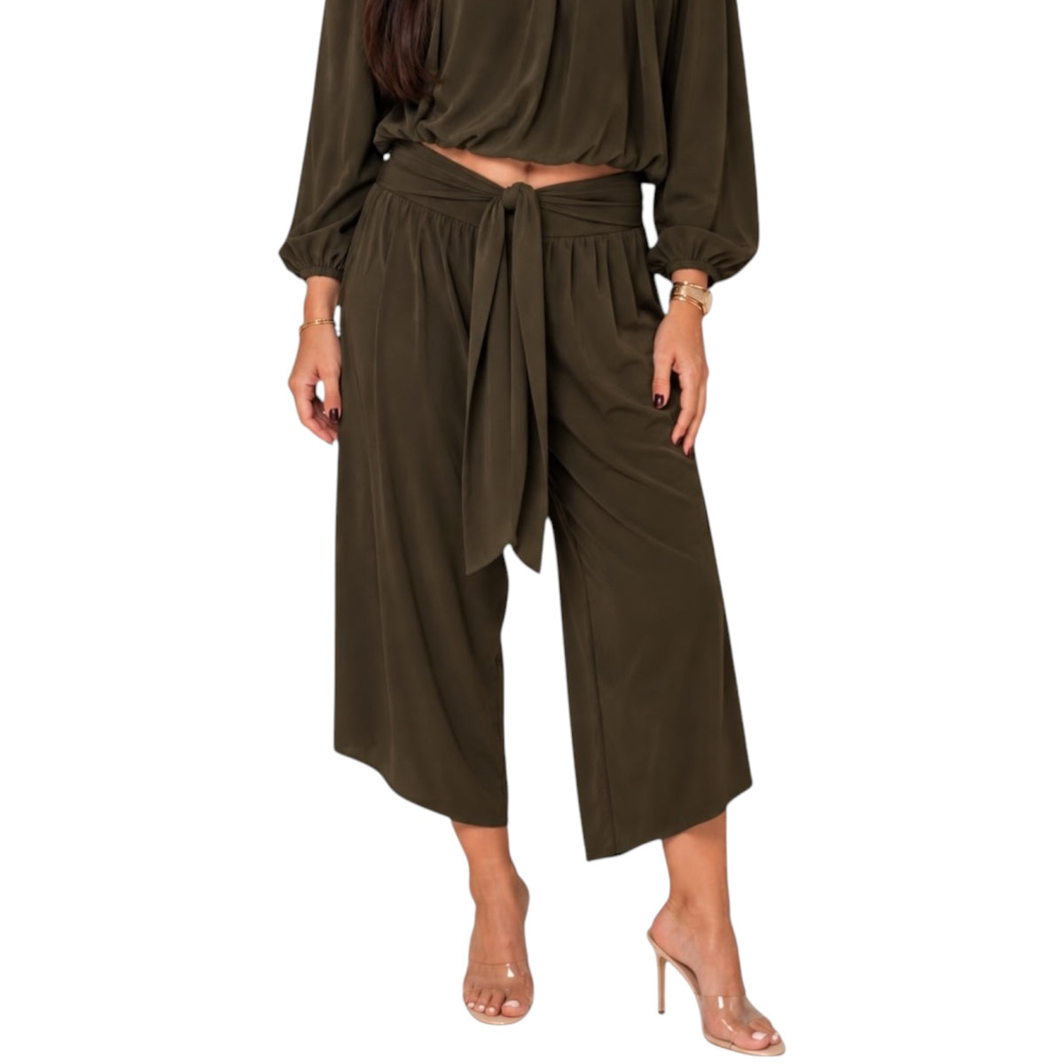 A model wearing an olive green two-piece set consisting of a long-sleeve crop top and matching wide-leg culotte pants with a tie waist, paired with clear high-heeled sandals against a white background.