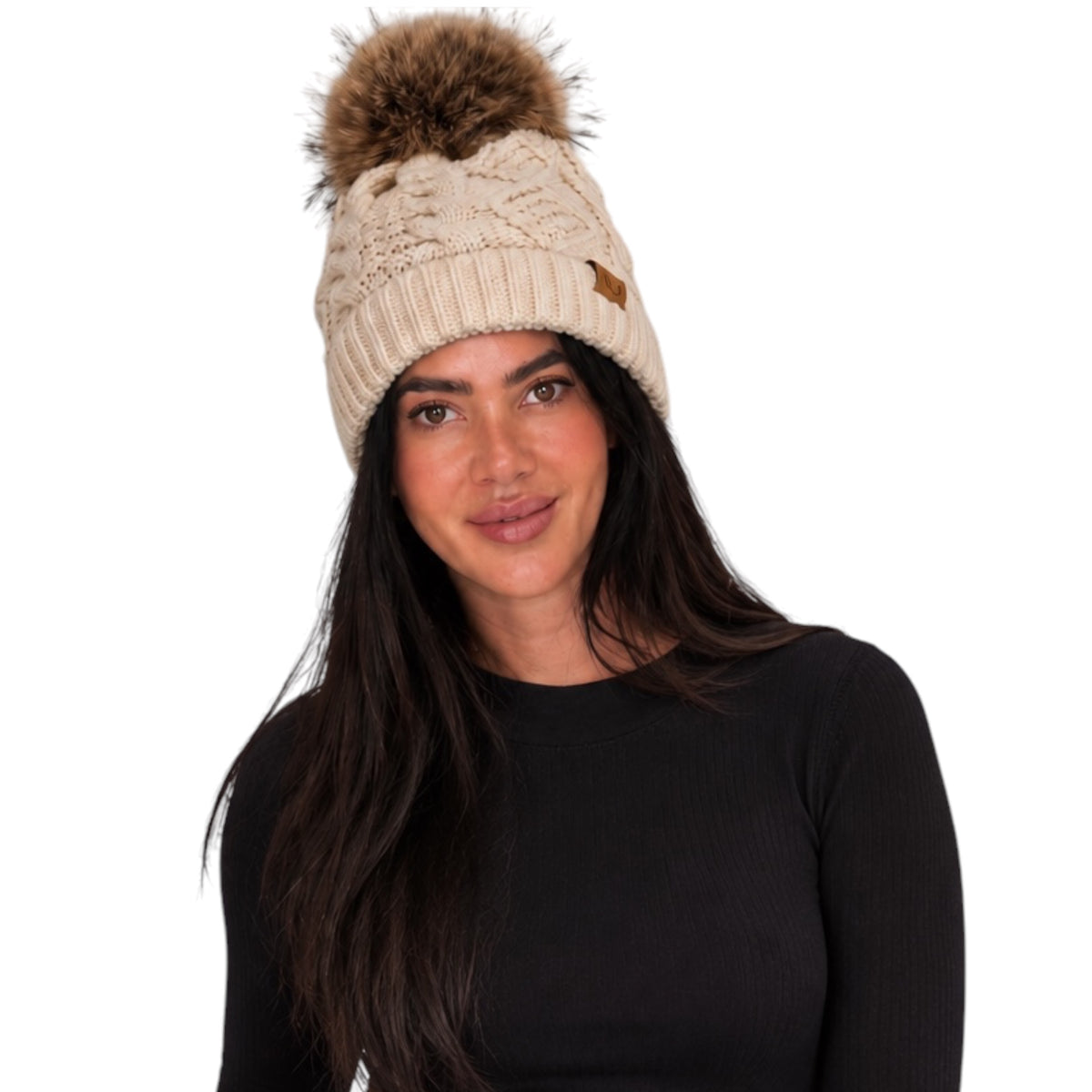 A woman wearing a beige cable-knit beanie with a large brown faux-fur pom-pom and a small brown leather logo patch, photographed against a plain white background.