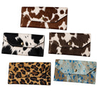 Five wallet-style clutch or bifold wallets with animal-print and speckled textures are arranged on a plain white background. The designs include brown and white cowhide, black and white cowhide, dark brown and white mottled, tan and black leopard, and tan and blue speckled.