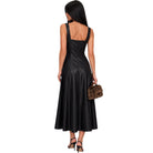 A woman shown from the back wearing a sleeveless black midi dress with a structured back and flared skirt, holding a small brown patterned handbag against a plain white background.