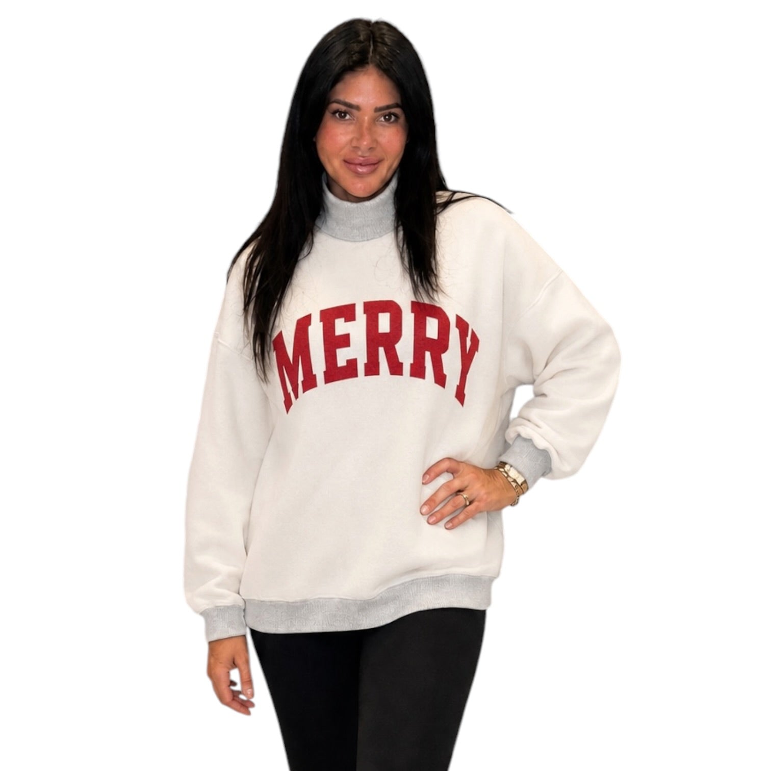 A woman modeling a cream-colored mock neck sweatshirt with 'MERRY' printed in red arched letters across the chest, paired with black pants, against a plain white background.