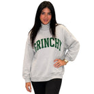 A woman modeling a light gray turtleneck sweatshirt with 'GRINCHY' printed in green arched letters across the chest, paired with black leggings, against a plain white background.
