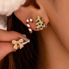 Close-up of a person wearing holiday-themed stud earrings, including a gold tree with green and blue stones and a candy cane, with another matching gold bow earring held near the ear.
