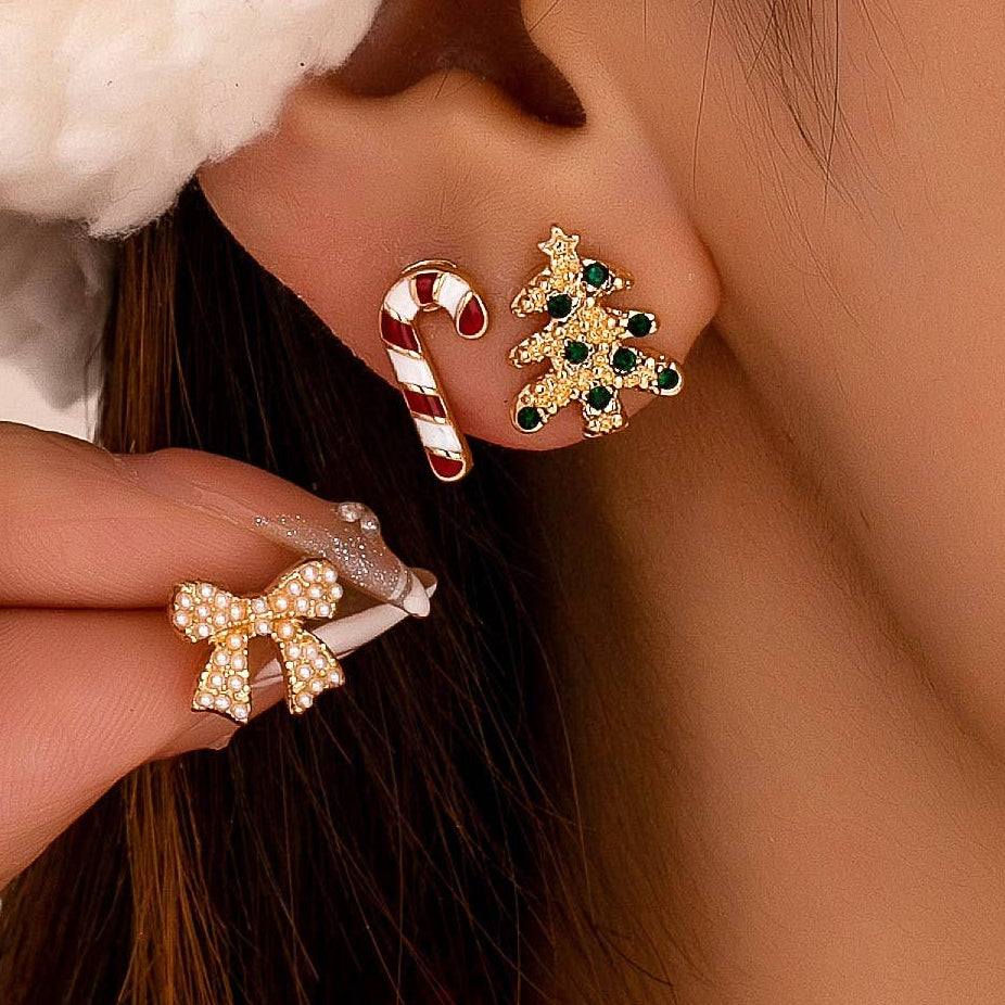 Close-up of a person wearing holiday-themed stud earrings, including a gold tree with green and blue stones and a candy cane, with another matching gold bow earring held near the ear.