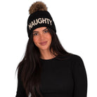 A woman wearing a black knit beanie with a brown faux-fur pom-pom and the word 'NAUGHTY' in white glittery letters across the front, against a plain white background.
