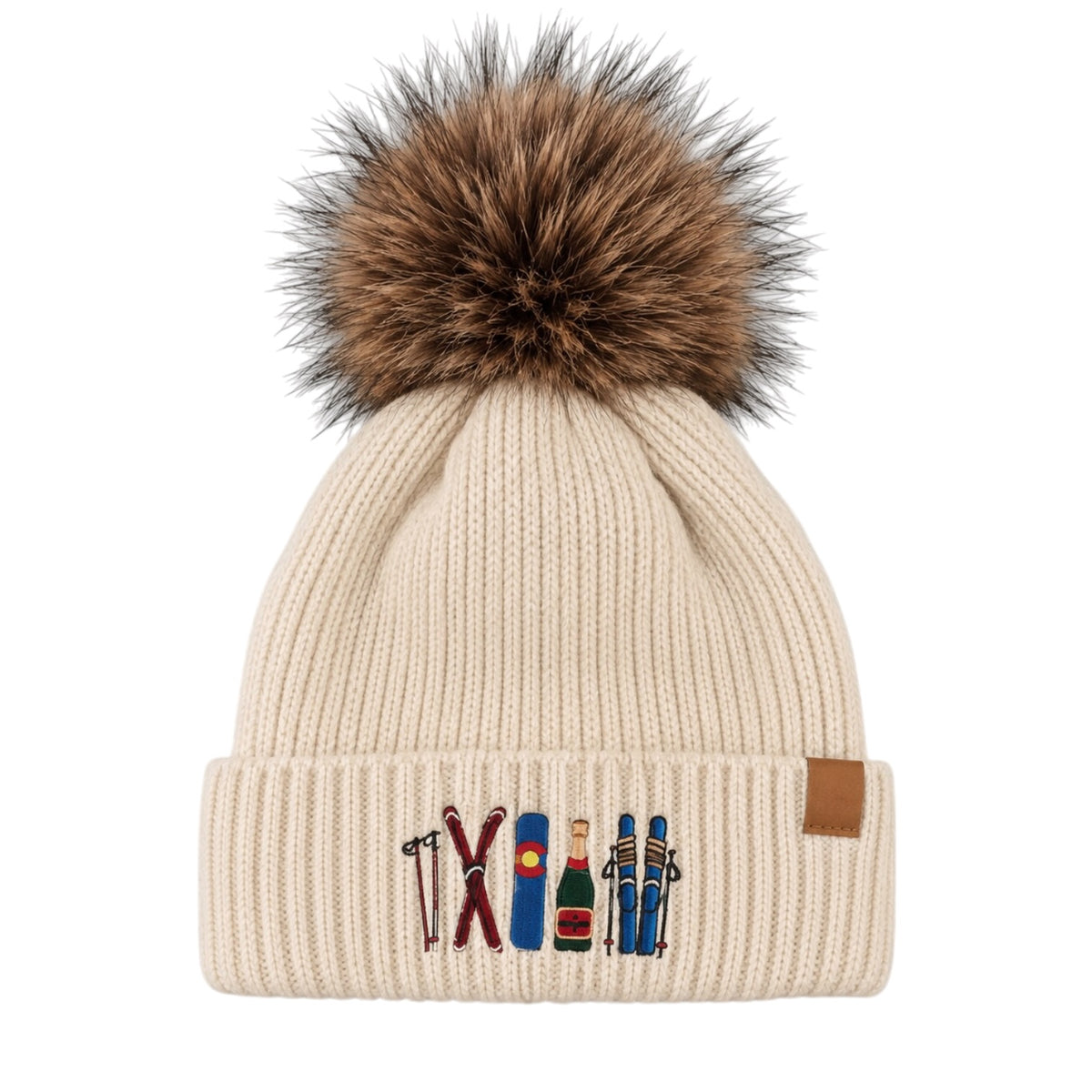 A cream-colored ribbed knit beanie hat with a large brown and black faux fur pom-pom on top. The front cuff features embroidered winter sports gear, including skis, a ski pole, a ski boot, a champagne bottle, and skiers, alongside a small brown leather brand tab on the side.