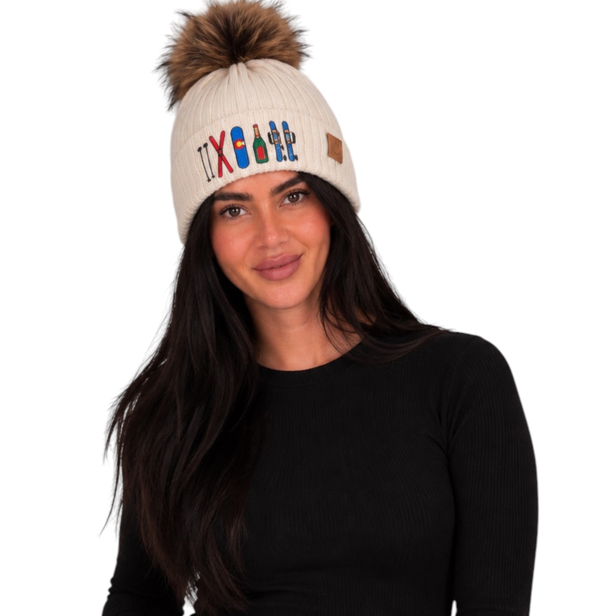 A woman wearing a cream ribbed knit beanie with a brown faux-fur pom-pom and colorful embroidered skis, paired with a black long-sleeve top, against a plain white background.