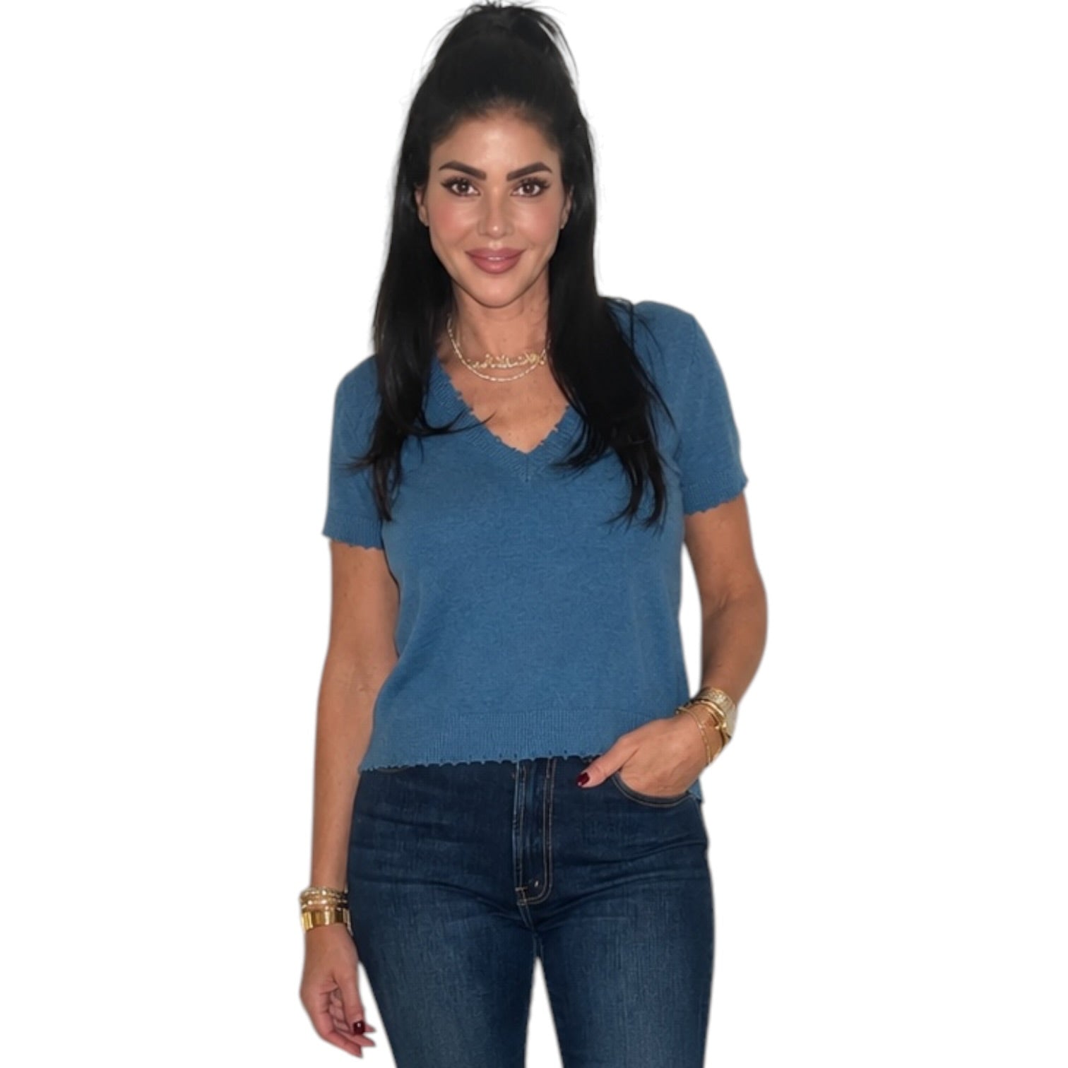 A woman modeling a short-sleeve blue V-neck knit top with a scalloped edge, paired with dark blue jeans, against a plain white background.