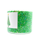 A clear glass jar candle filled with green glitter and small multicolored confetti, with a white paper label showing a Christmas tree illustration, on a plain white background.