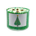 A three-wick scented candle in a clear glass jar with a green glittery exterior and a white front label featuring a green Christmas tree illustration. The lid is decorated with green sprinkles and a gold star, with three dark wicks visible.