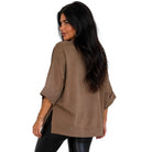 A woman with long dark hair is shown from the back wearing an oversized taupe-brown short-sleeve top and black pants against a plain white background.