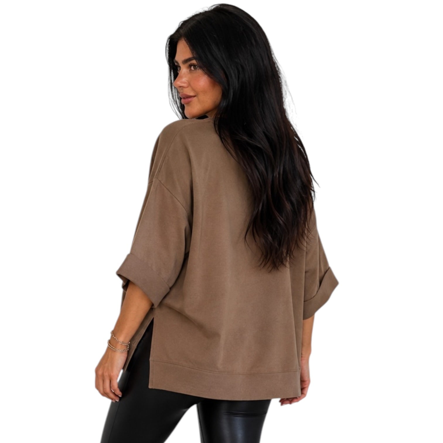 A woman with long dark hair is shown from the back wearing an oversized taupe-brown short-sleeve top and black pants against a plain white background.