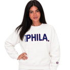 A woman modeling a white crewneck sweatshirt with 'PHILA.' printed across the chest in navy blue with a red outline, and a small circular logo on the left sleeve.