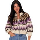 A woman wearing a cropped knit zip-up cardigan with a cream base and brown, black, and purple geometric patterns, paired with blue jeans against a plain white background.