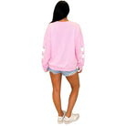 A woman shown from the back wearing a light pink crewneck sweatshirt with small white cartoon graphics on the sleeves, paired with denim shorts and white sneakers, against a plain white background.