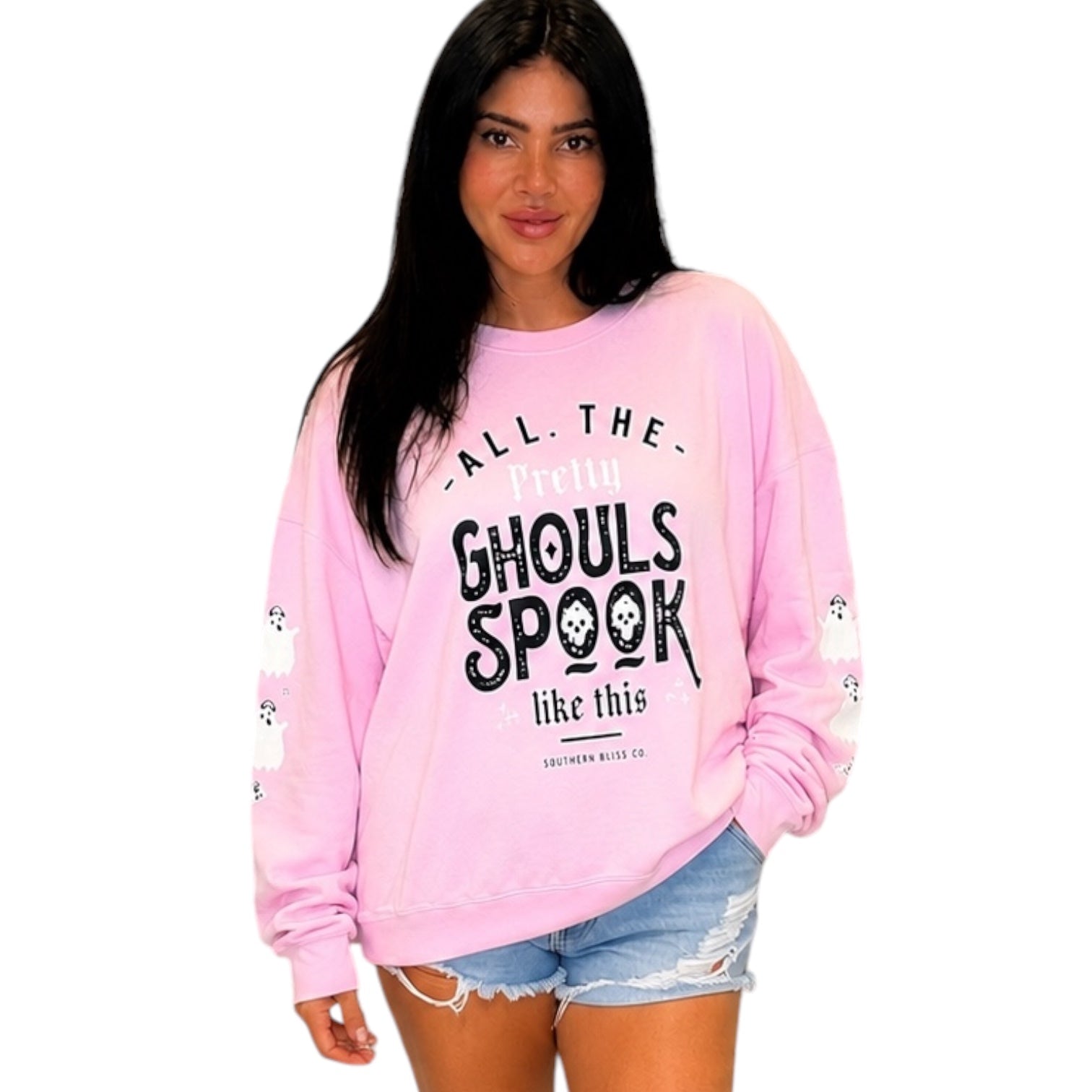 A woman modeling a light pink crewneck sweatshirt with a black graphic text print that reads 'ALL THE PRETTY GHOULS SPOOK like this' and small white skull details on the sleeves, paired with light blue denim shorts against a plain white background.