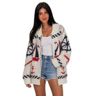 A woman modeling a cream-colored knit cardigan with nautical patterns including sailboats, anchors, and compasses in navy blue and red, styled with a white tank top and denim shorts against a plain white background.