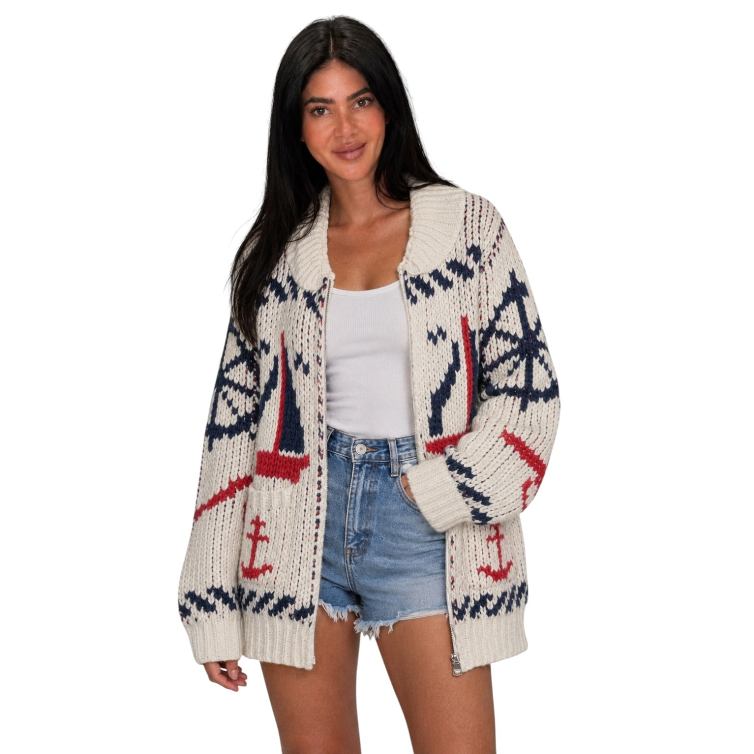 A woman modeling a cream-colored knit cardigan with nautical patterns including sailboats, anchors, and compasses in navy blue and red, styled with a white tank top and denim shorts against a plain white background.