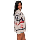 A woman modeling a chunky knit cardigan with a nautical theme, featuring red and navy blue graphics of a sailboat, anchor, and ship's wheel on an off-white background.