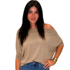 A woman with long dark hair wearing a beige off-the-shoulder knit top and blue jeans, standing against a plain white background.