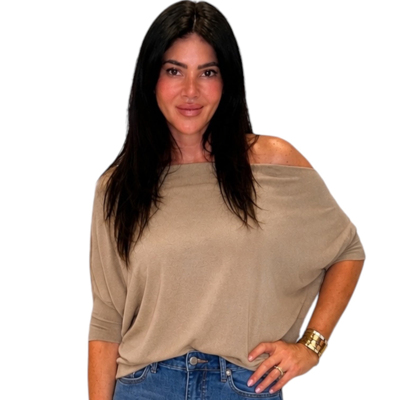 A woman with long dark hair wearing a beige off-the-shoulder knit top and blue jeans, standing against a plain white background.