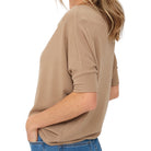 A woman wearing a loose, short-sleeve knit top in a warm taupe-beige color, paired with blue jeans, shown from a side-back angle against a plain white background.