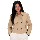 A woman modeling a cropped beige double-breasted jacket with black buttons, paired with white trousers, against a solid white background.