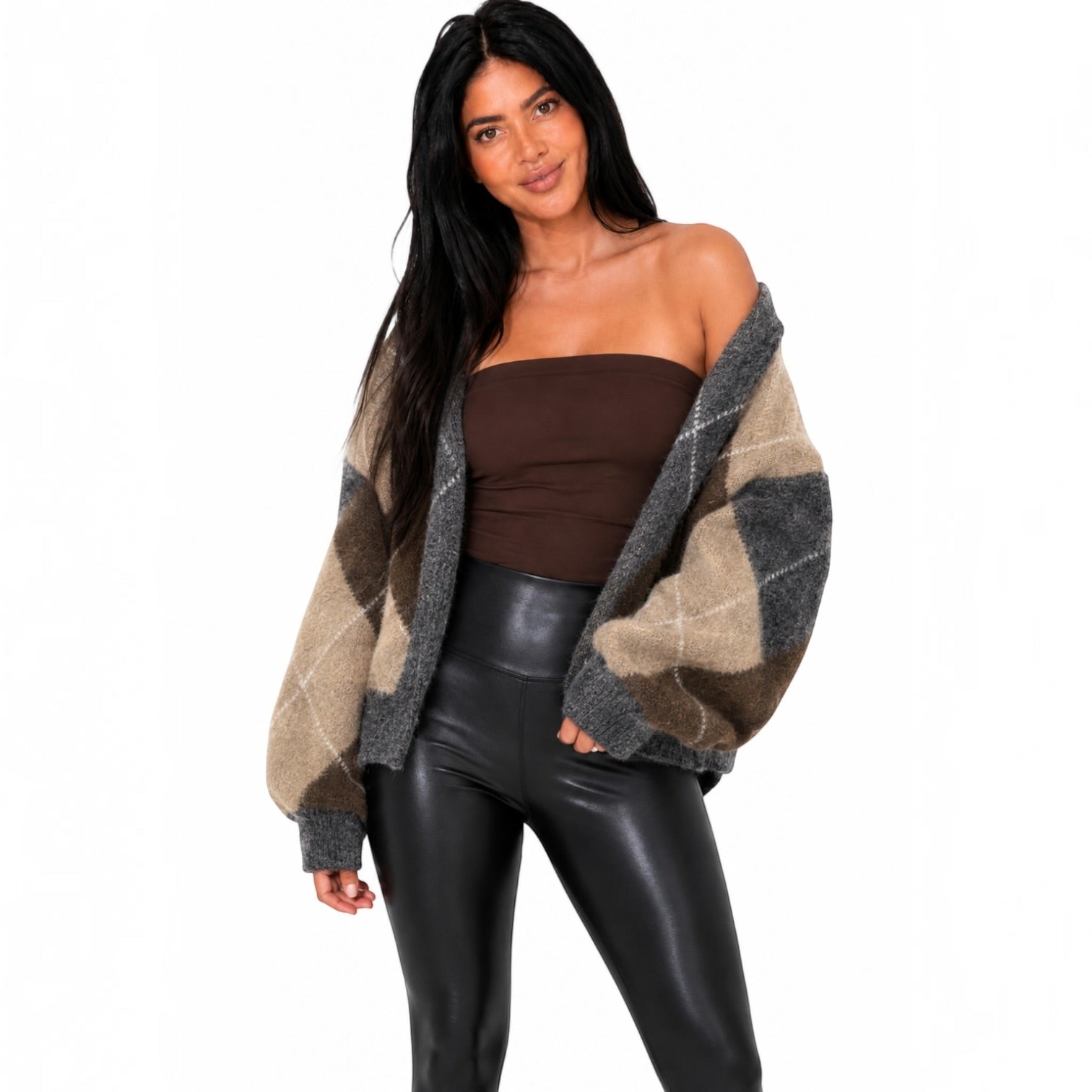 A woman modeling a chunky knit cardigan with a brown, beige, and gray argyle pattern, worn open over a brown strapless top and black faux-leather pants, against a plain white background.