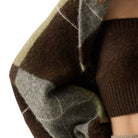 Close-up of a person wearing a chunky knit sweater with a brown, gray, and muted olive plaid pattern, paired with a solid dark brown ribbed knit sleeve or undershirt.