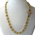 A gold-toned link necklace displayed on a white fabric mannequin bust. The necklace features textured oval links and a decorative cross-shaped charm embellished with pink, purple, and dark red faceted gemstones.