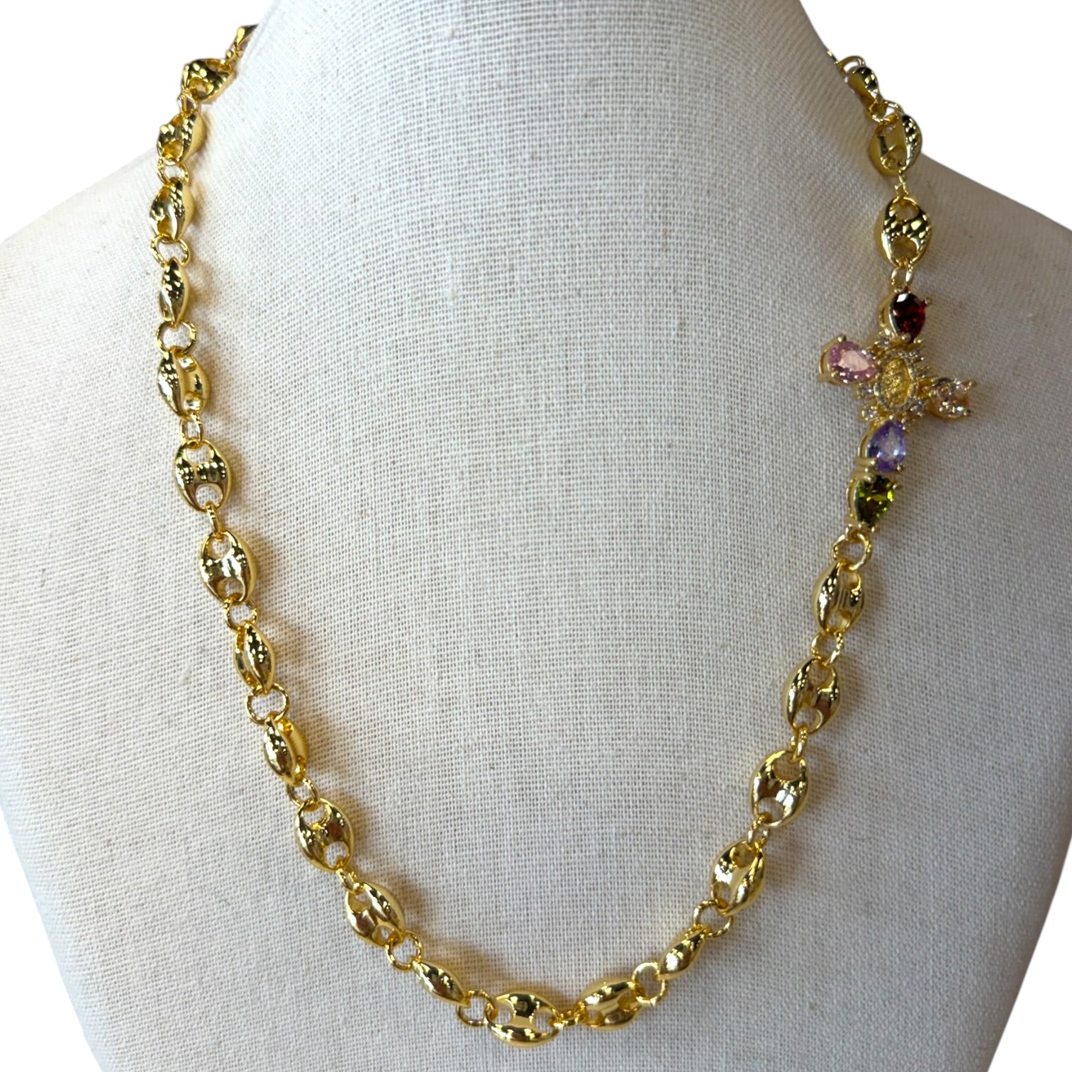 A gold-toned link necklace displayed on a white fabric mannequin bust. The necklace features textured oval links and a decorative cross-shaped charm embellished with pink, purple, and dark red faceted gemstones.