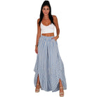 A woman modeling a two-piece outfit consisting of a white crop top and high-waisted, wide-leg blue and white striped maxi pants with side slits, set against a plain white background.