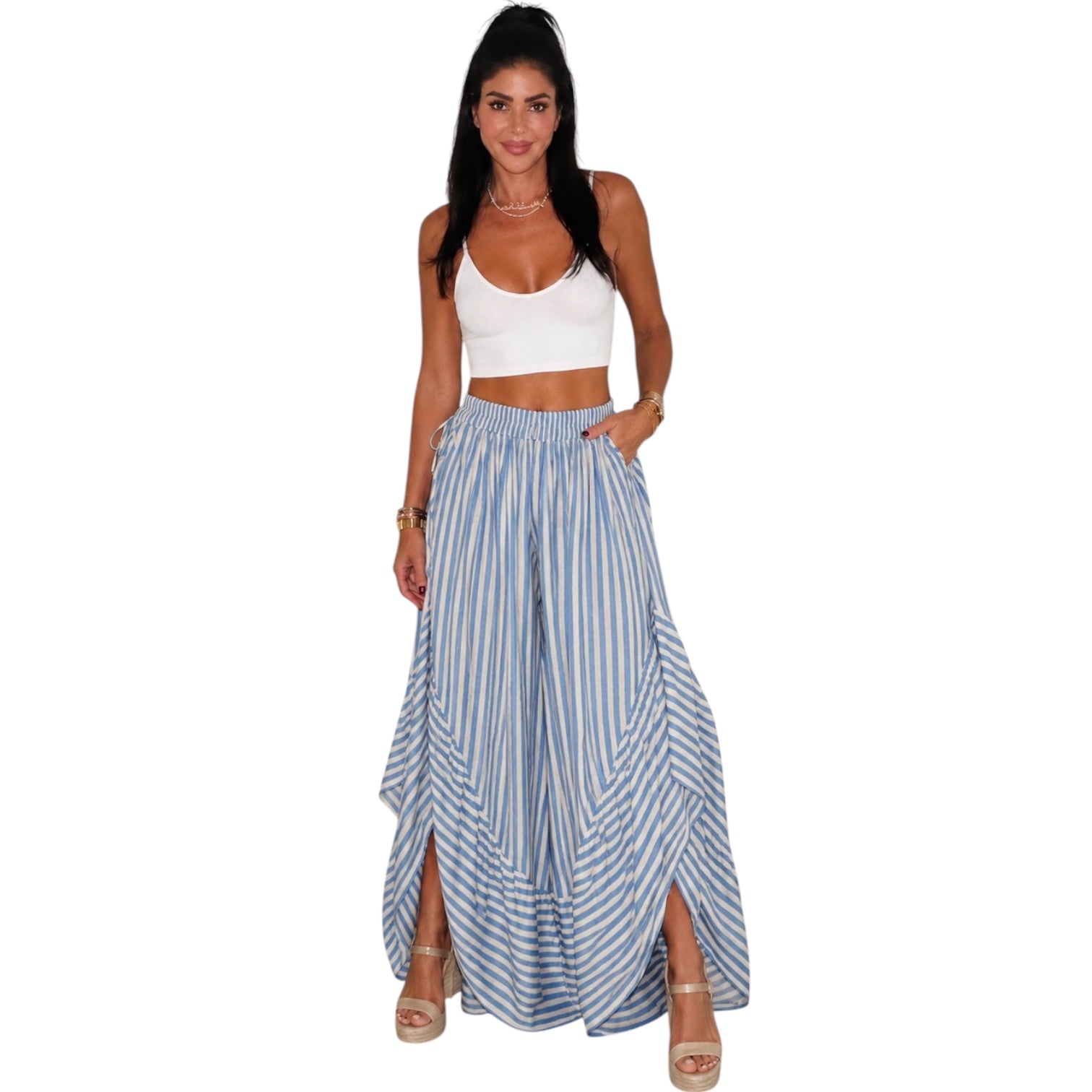 A woman modeling a two-piece outfit consisting of a white crop top and high-waisted, wide-leg blue and white striped maxi pants with side slits, set against a plain white background.