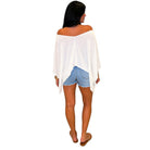 A woman shown from the back wearing a white off-the-shoulder poncho top, light blue denim shorts, and brown flat sandals against a plain white background.