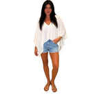 A woman modeling a white lightweight draped long-sleeve top paired with light blue denim shorts and brown sandals, set against a plain white background.