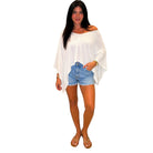 A woman modeling a white off-the-shoulder long-sleeve top paired with light blue denim shorts and brown sandals, set against a plain white background.