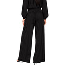 A model shown from the back wearing a black long-sleeve jumpsuit with balloon sleeves and wide-leg trousers, paired with black high-heeled sandals against a plain white background.