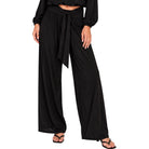 A model wearing black wide-leg draped pants with a matching waist tie, paired with black sandals against a plain white background.