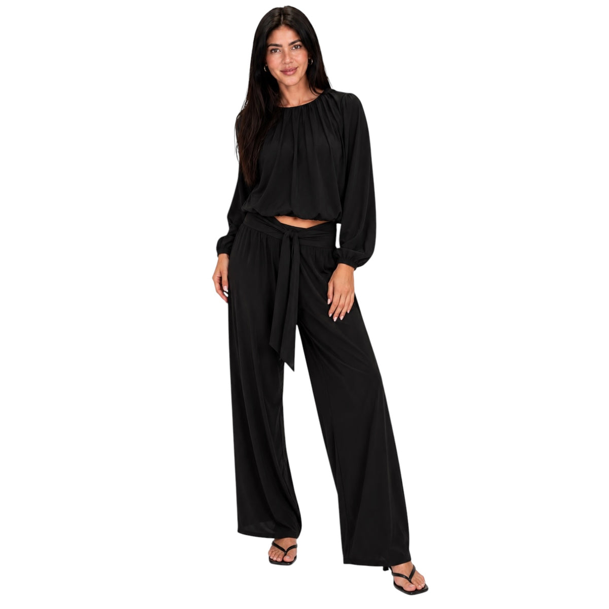 A woman modeling a black two-piece outfit consisting of a long-sleeve cropped top and matching wide-leg pants with a tie waist, paired with black sandals against a plain white background.