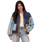A woman modeling a patchwork bandana quilted jacket with navy and light blue patterned panels and tan trim, worn over a white top and light-wash jeans against a plain white background.
