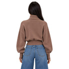 A woman viewed from the back wearing a cropped brown knit sweater with a high ribbed turtleneck and blue jeans, against a plain white background.