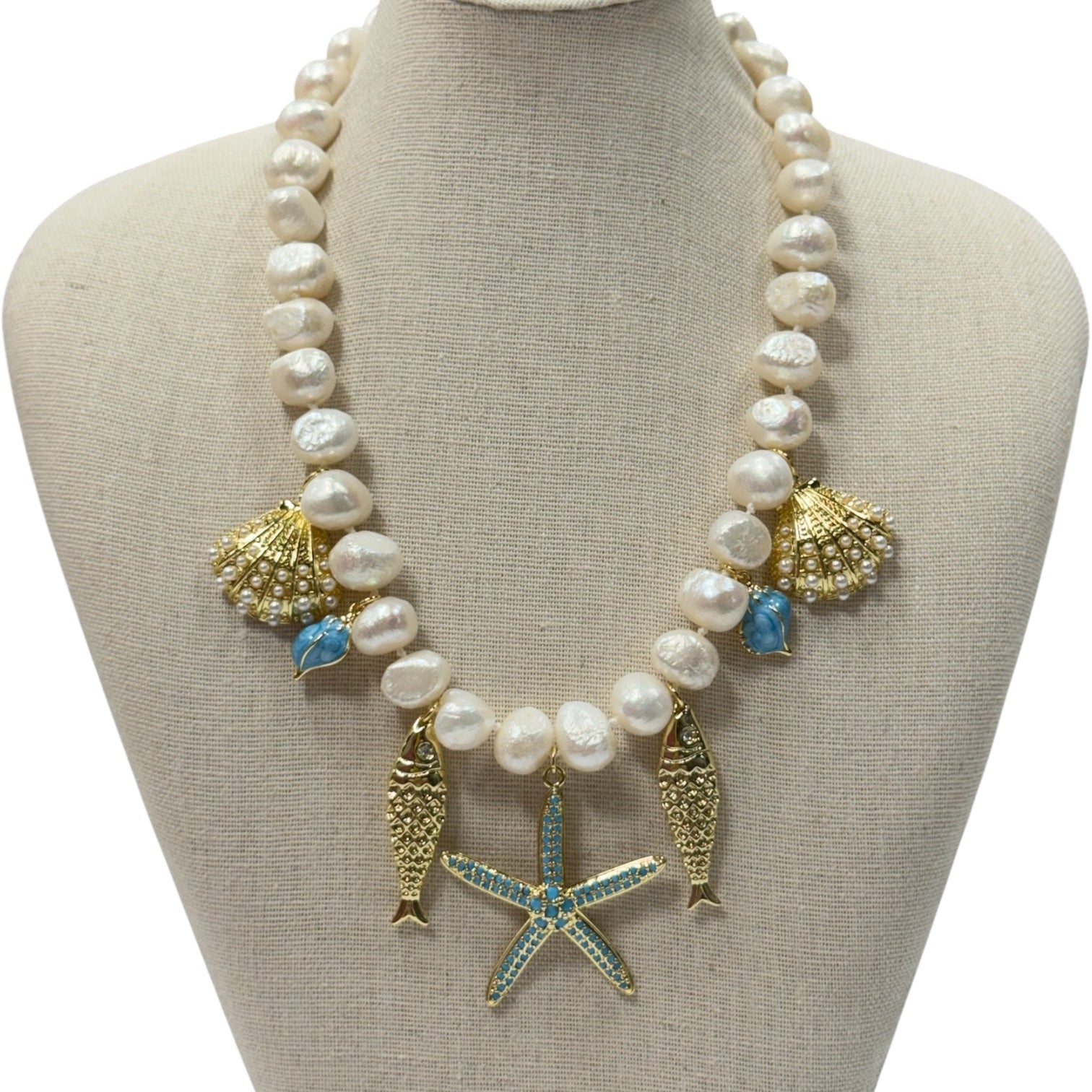 A pearl and gold statement necklace with seashell, starfish, and fish charms, displayed on a beige fabric jewelry bust.