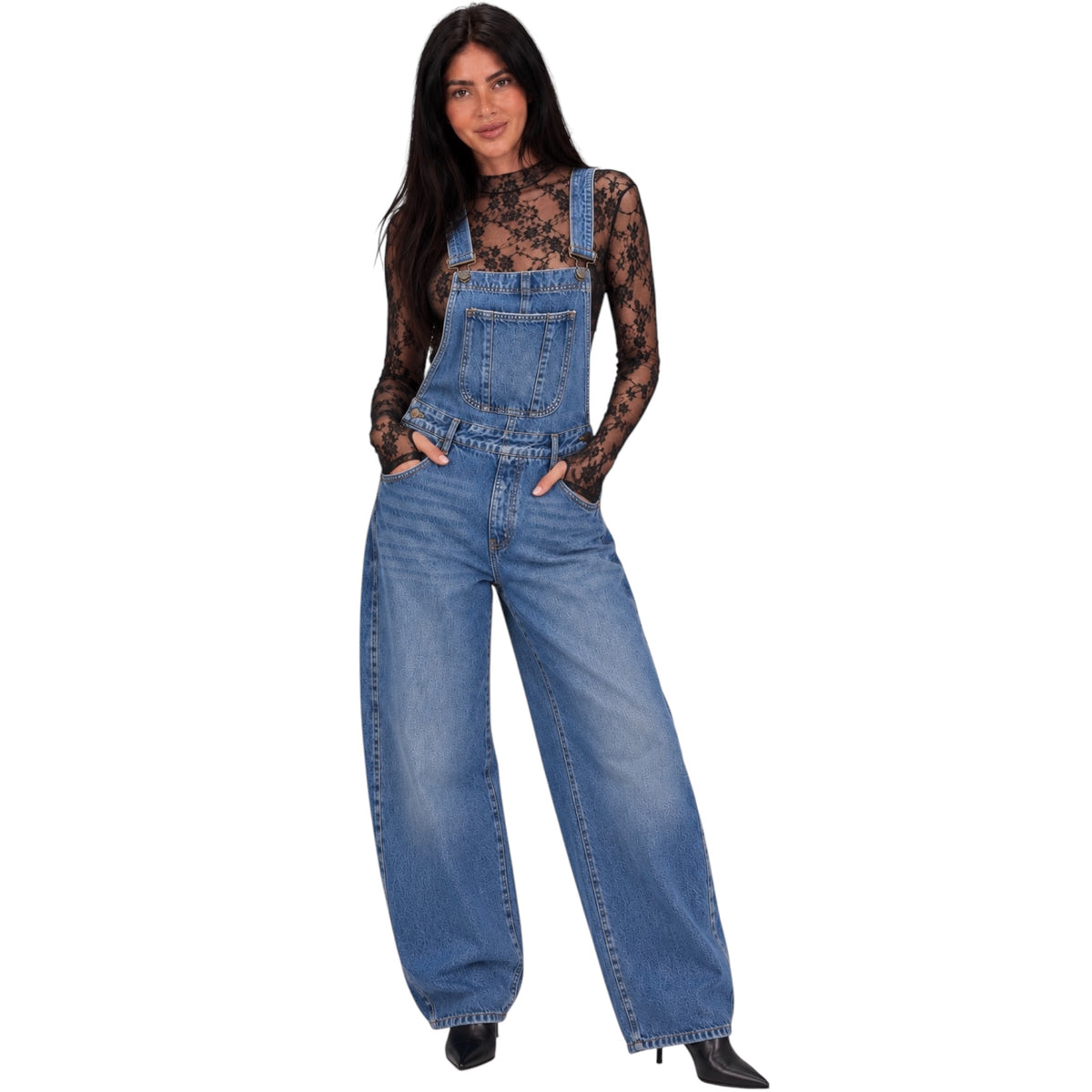 A woman modeling a pair of wide-leg blue denim overalls worn over a sheer black lace long-sleeve top, set against a plain white background.
