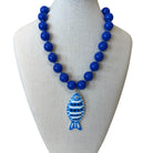 A beaded necklace displayed on a cream fabric mannequin bust. The necklace features glossy cobalt blue round beads and a central fish-shaped pendant with blue and white stripes and a blue eye.