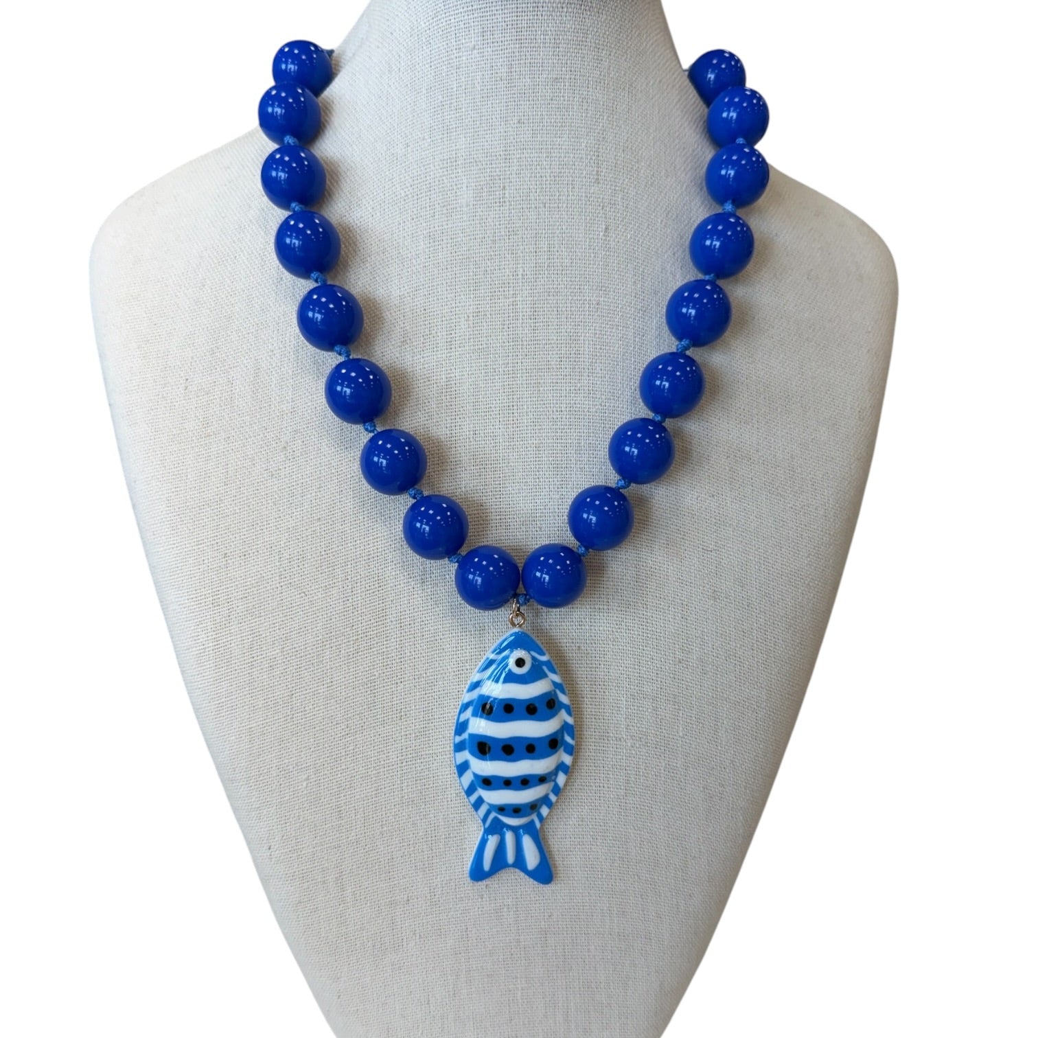 A beaded necklace displayed on a cream fabric mannequin bust. The necklace features glossy cobalt blue round beads and a central fish-shaped pendant with blue and white stripes and a blue eye.