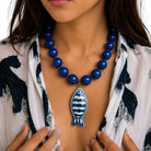 A close-up of a woman wearing a blue beaded necklace with a ceramic fish pendant, styled with a white and navy patterned shirt.