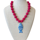 A bright pink beaded necklace displayed on a cream fabric jewelry bust. The necklace features glossy hot pink round beads with a central pendant shaped like a blue and white striped fish.